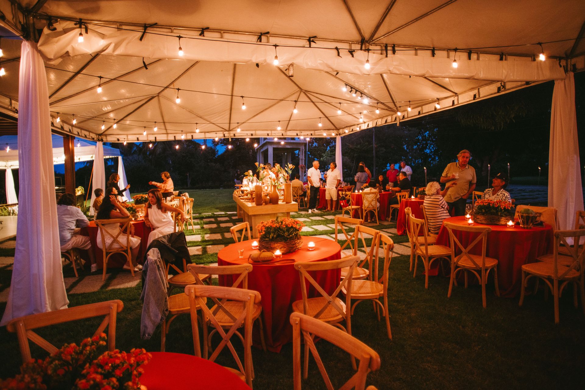 Charming outdoor evening event setup with string lights, red tablecloths, and wooden chairs under a canopy.